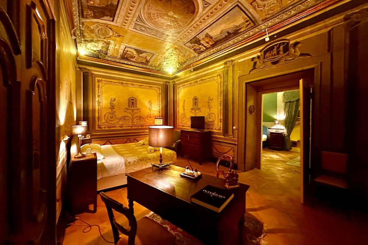 Ein luxuriöses Hotelzimmer mit aufwendiger Deckenmalerei, einem Bett mit weißer Bettdecke, einem Schreibtisch mit dem Buch 'INTERIORS' und einem Sessel.
