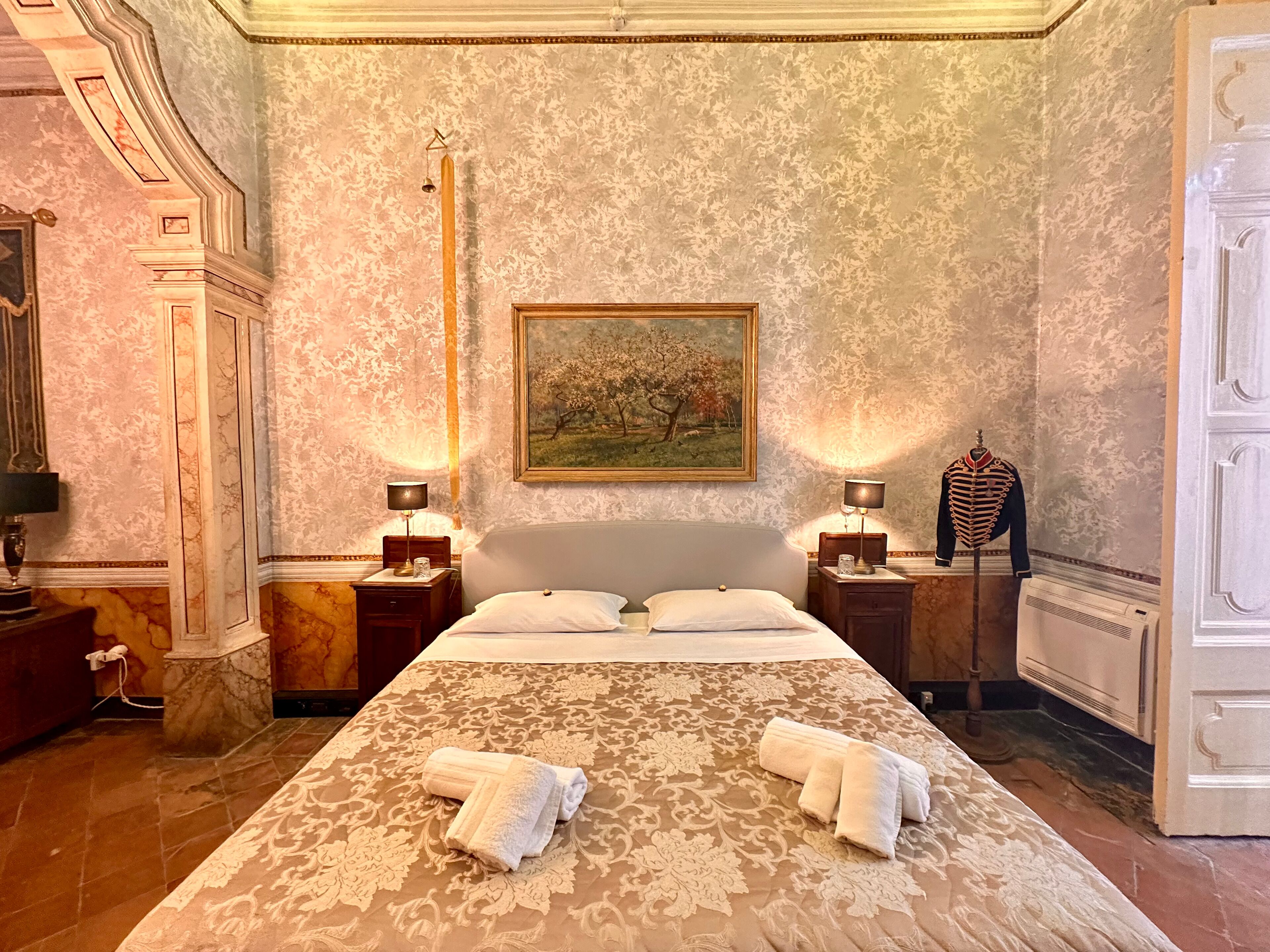Quarto de hotel com uma cama grande, duas mesas de cabeceira com luminárias, um quadro na parede e um cabideiro com um casaco.