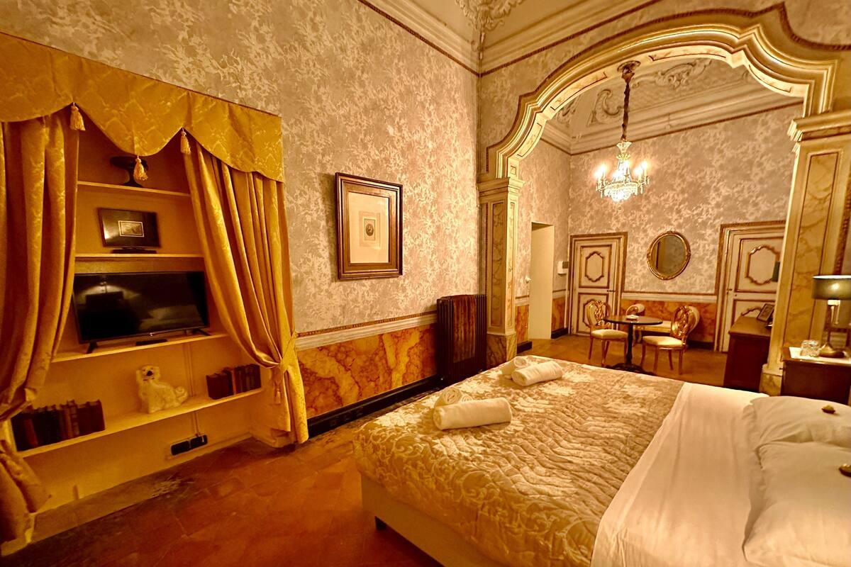 Ein Hotelzimmer mit einem großen Bett, einem Fernseher auf einem Regal, einer Kandelaber und einem Essbereich mit Stühlen und einem Tisch.