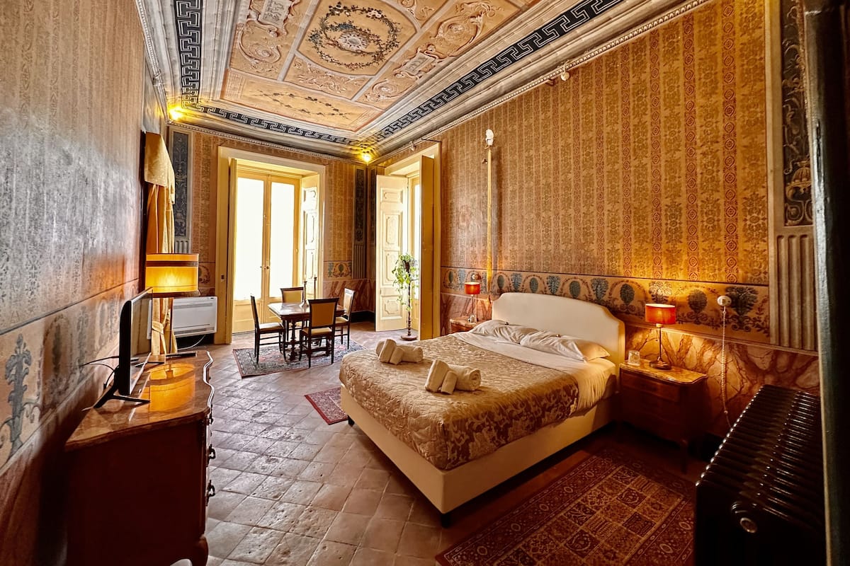 Ein luxuriöses Schlafzimmer mit einem großen Bett, einem Schreibtisch und einem Sessel, verziert mit aufwendigen Verzierungen an Decke und Wand.