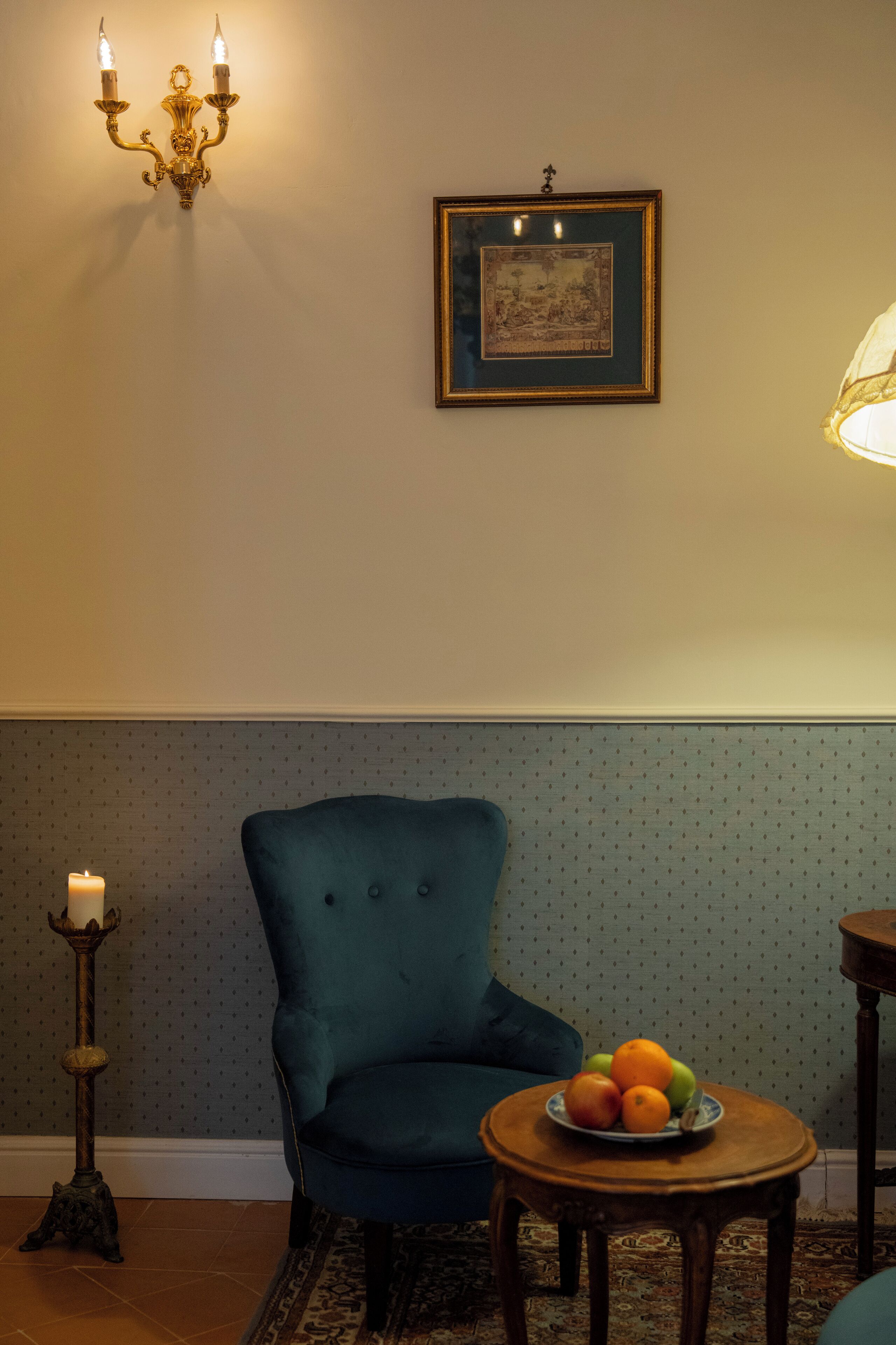 Un sillón color turquesa, una mesita redonda con fruta, un cuadro colgado en la pared y un candelabro con una vela.