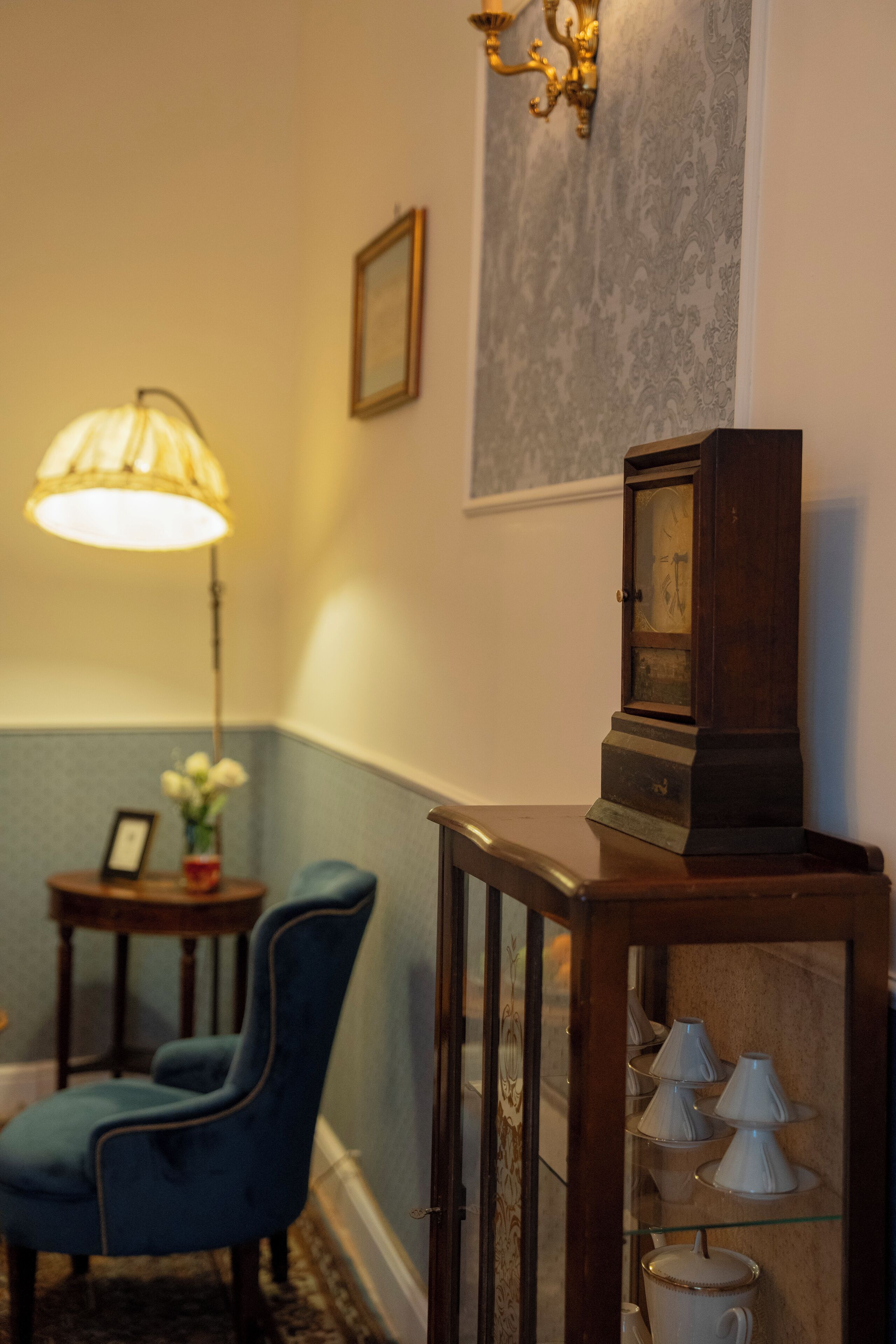 A room with a blue armchair, a wooden side table with a clock, a lamp, and a wall-mounted picture frame.