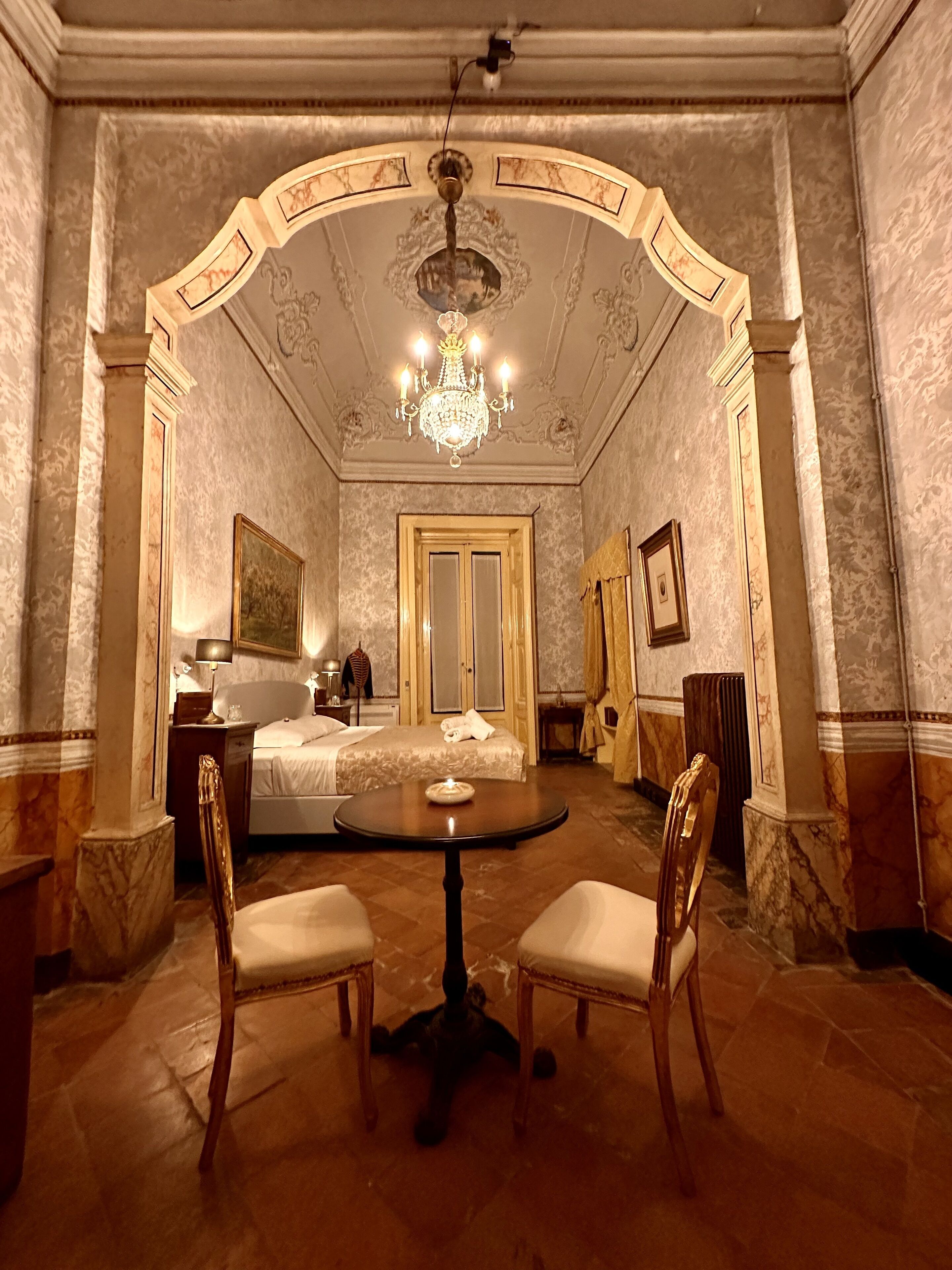 A luxurious hotel room with a marble floor, a chandelier, a bed with a canopy, a round table with chairs, and a large mirror.