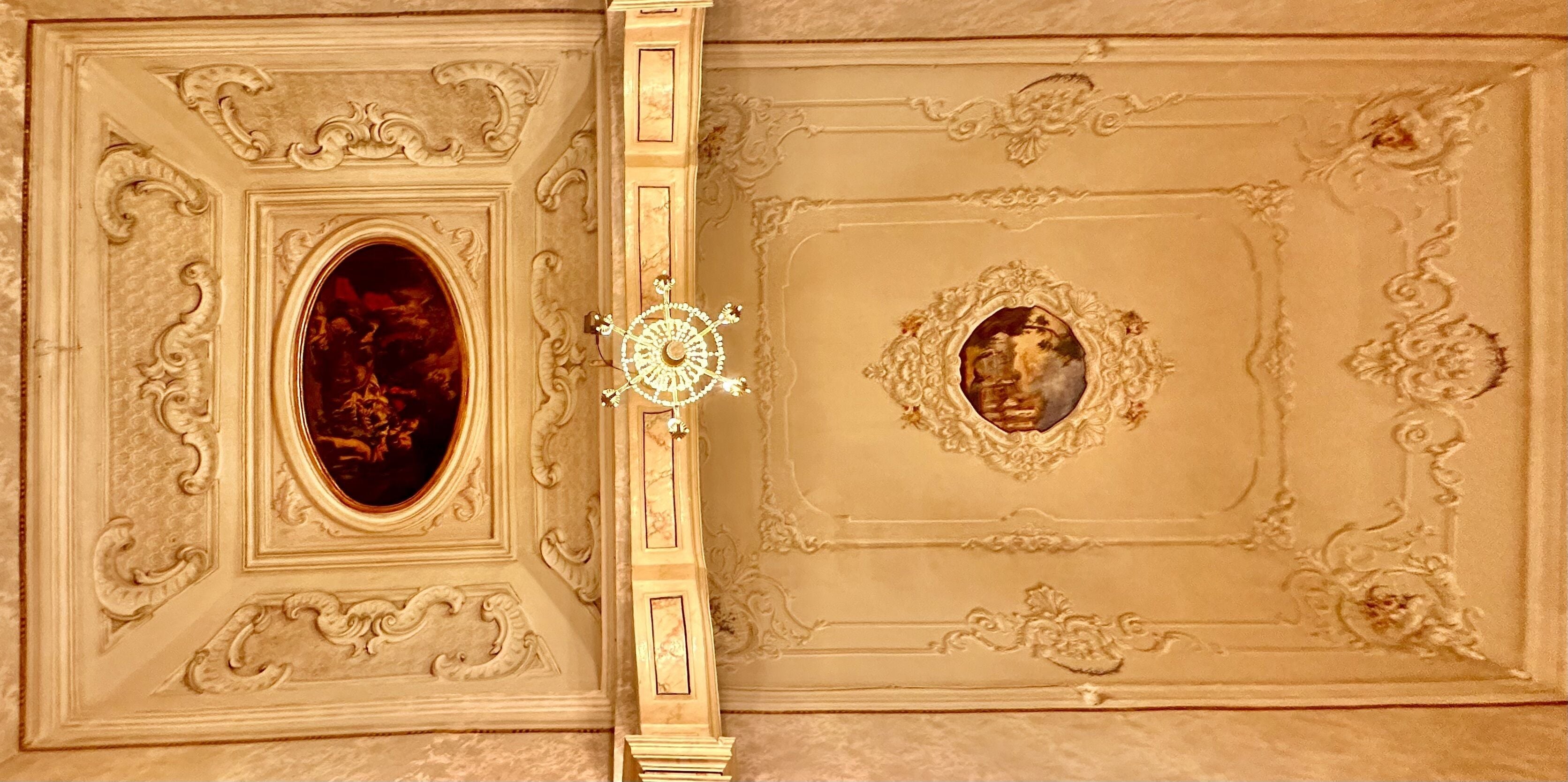 A ceiling with ornate plasterwork, a central chandelier, and two framed paintings.