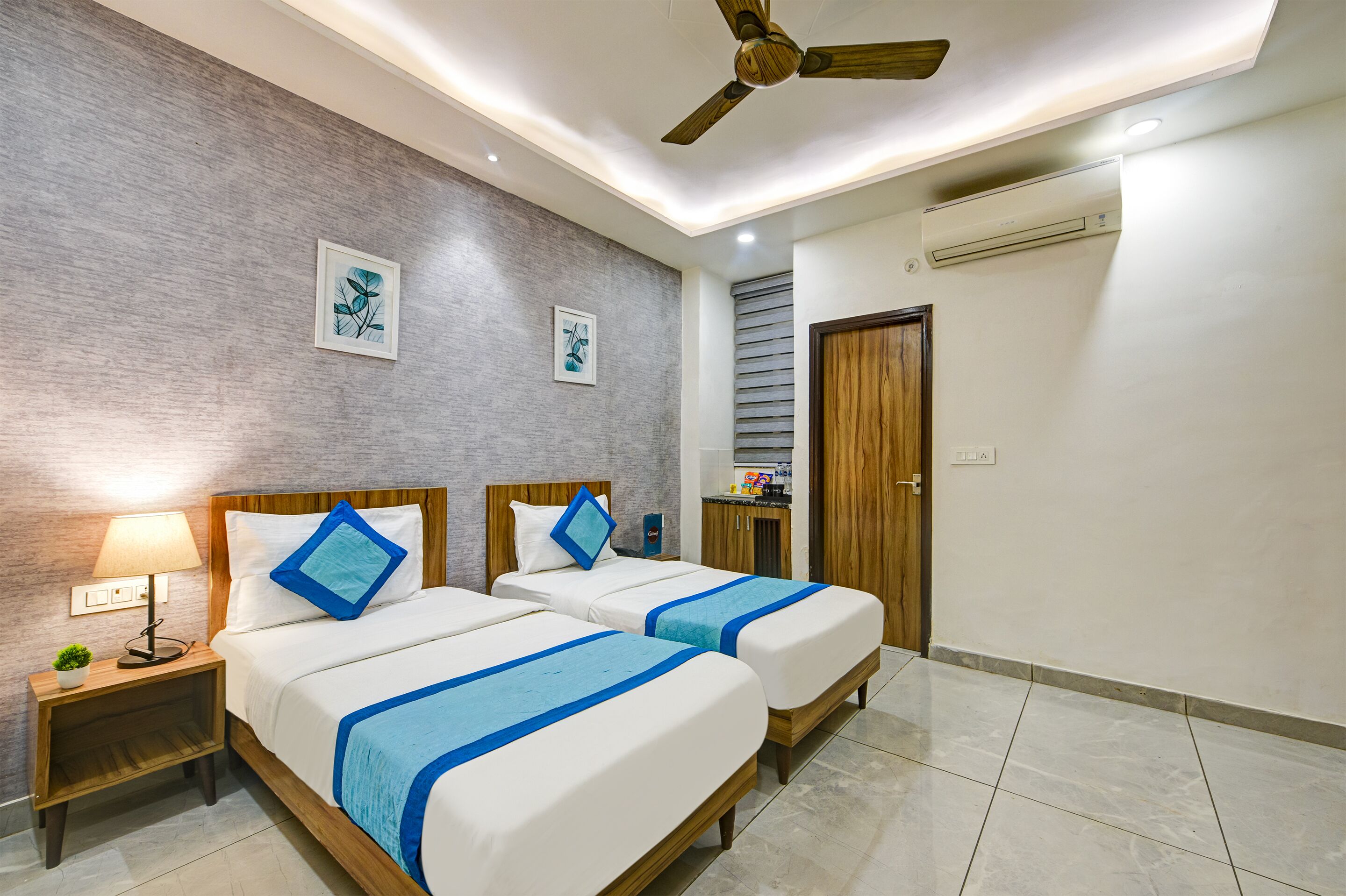 A hotel room with two beds, a ceiling fan, a wall-mounted air conditioner, and a small kitchenette.