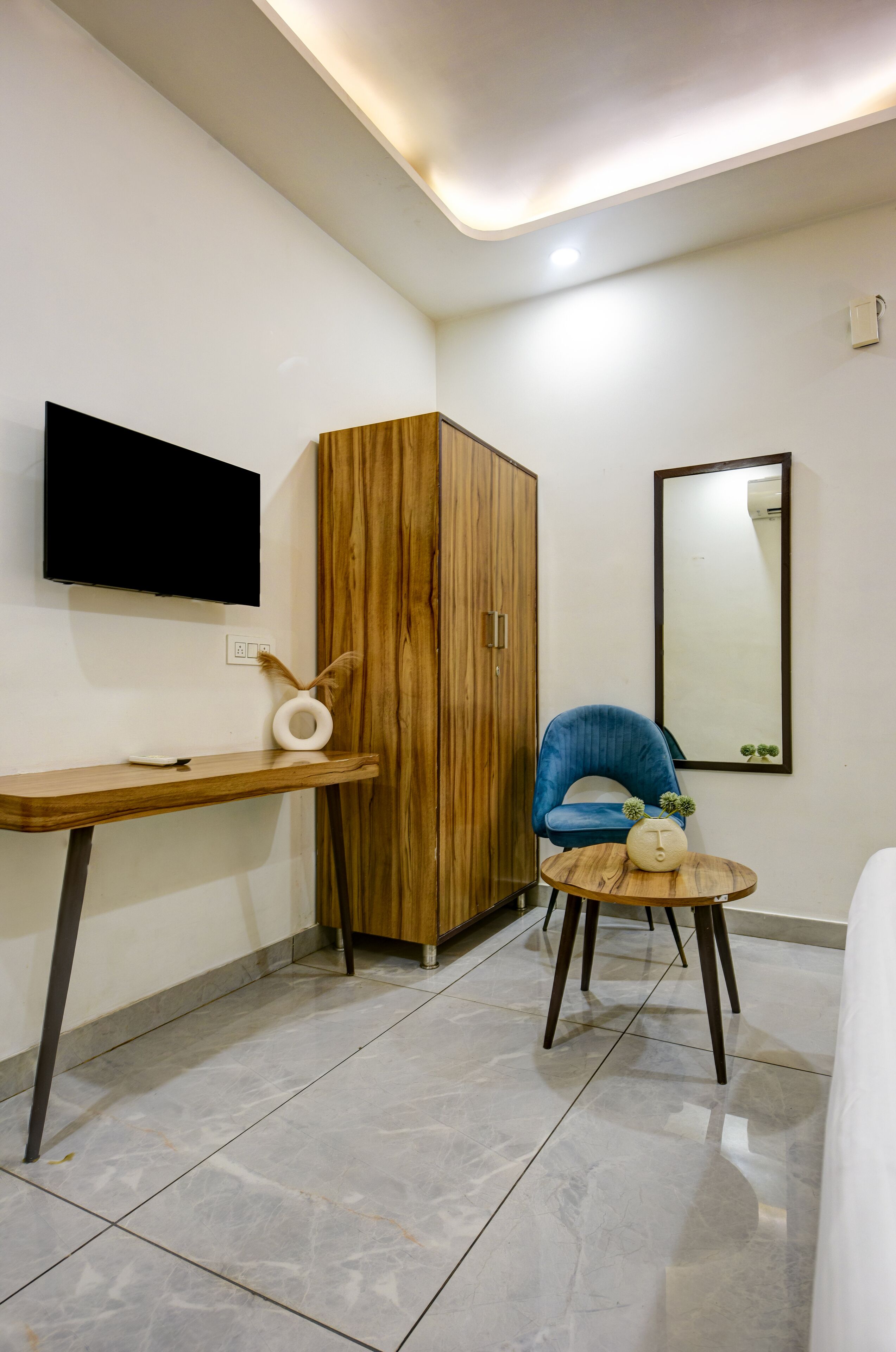 Een moderne kamer met een houten garderobe, een blauwe stoel, een klein rond tafeltje, een flatscreen televisie aan de muur en een spiegel.
