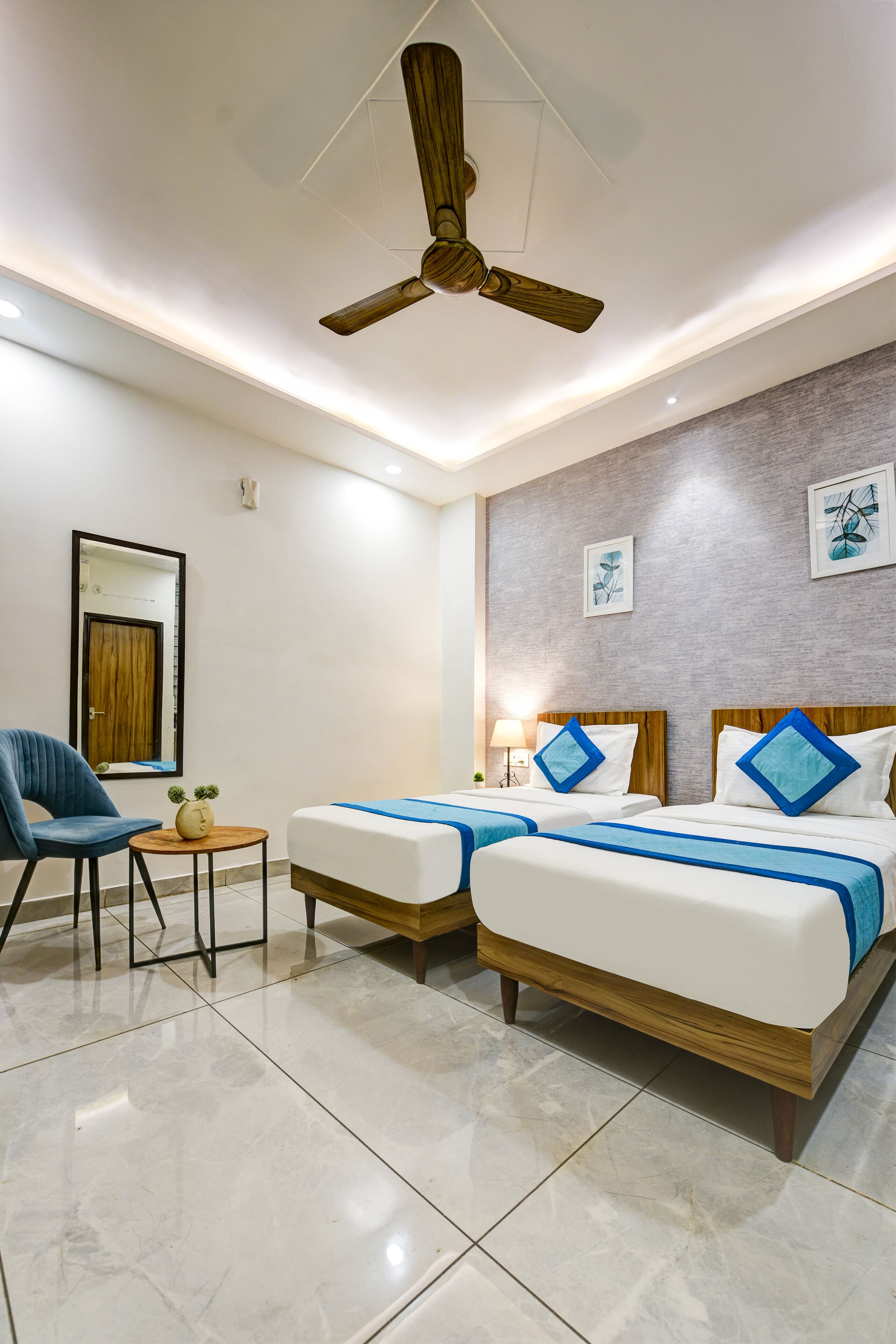 Une chambre d’hôtel avec deux lits, un ventilateur de plafond, une petite table avec une chaise et des œuvres d’art accrochées aux murs.