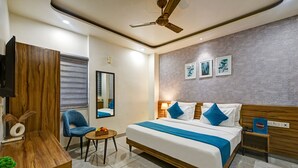 A modern hotel room with a large bed, a ceiling fan, a small table with fruit, a blue chair, and artwork on the wall. - Gallivanto Inn (New Delhi)
