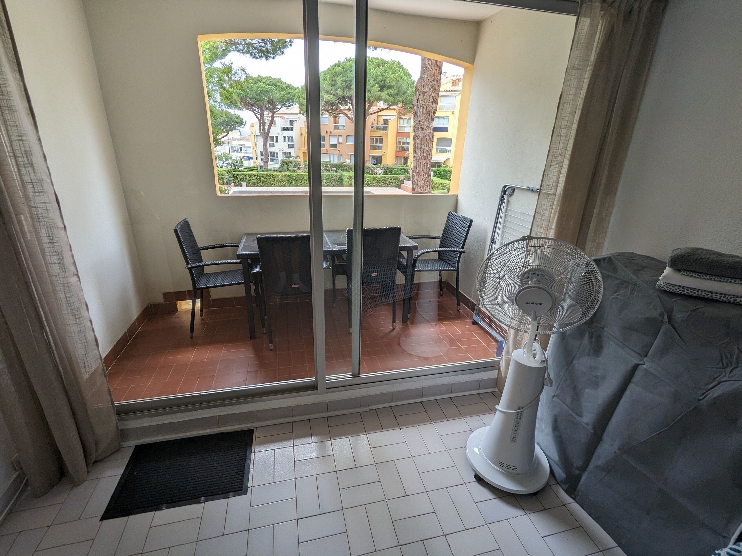 A balcony with a table and chairs, a fan, and a view of buildings and trees.