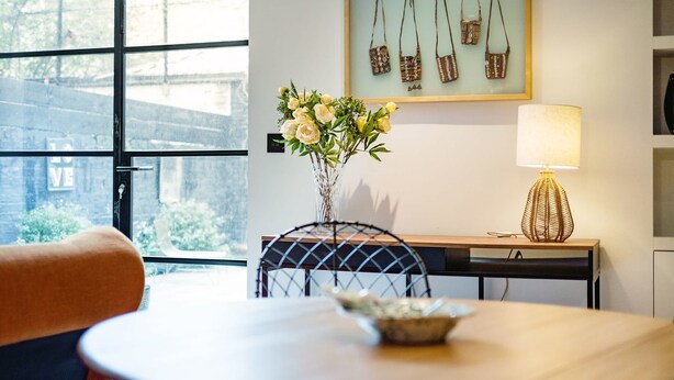 Un comedor con una mesa, sillas, un florero con flores, una lámpara y una pared con bolsos colgando.