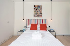 Une chambre d’hôtel moderne avec un grand lit, une tête de lit à rayures, deux tables de chevet, une œuvre d’art colorée inspirée des bandes dessinées et un seul luminaire suspendu.