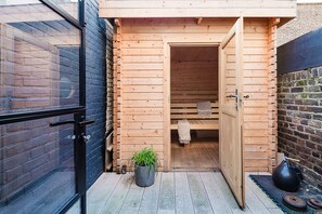 A wooden sauna with a bench and a door, a potted plant, and a sauna mat on the floor.
