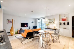 A modern living room with a sofa, dining table, and chairs, a flat-screen TV, and a large window. - Notting Hill 2 Bed with Sauna - by Out of Office Lifestyle (London)