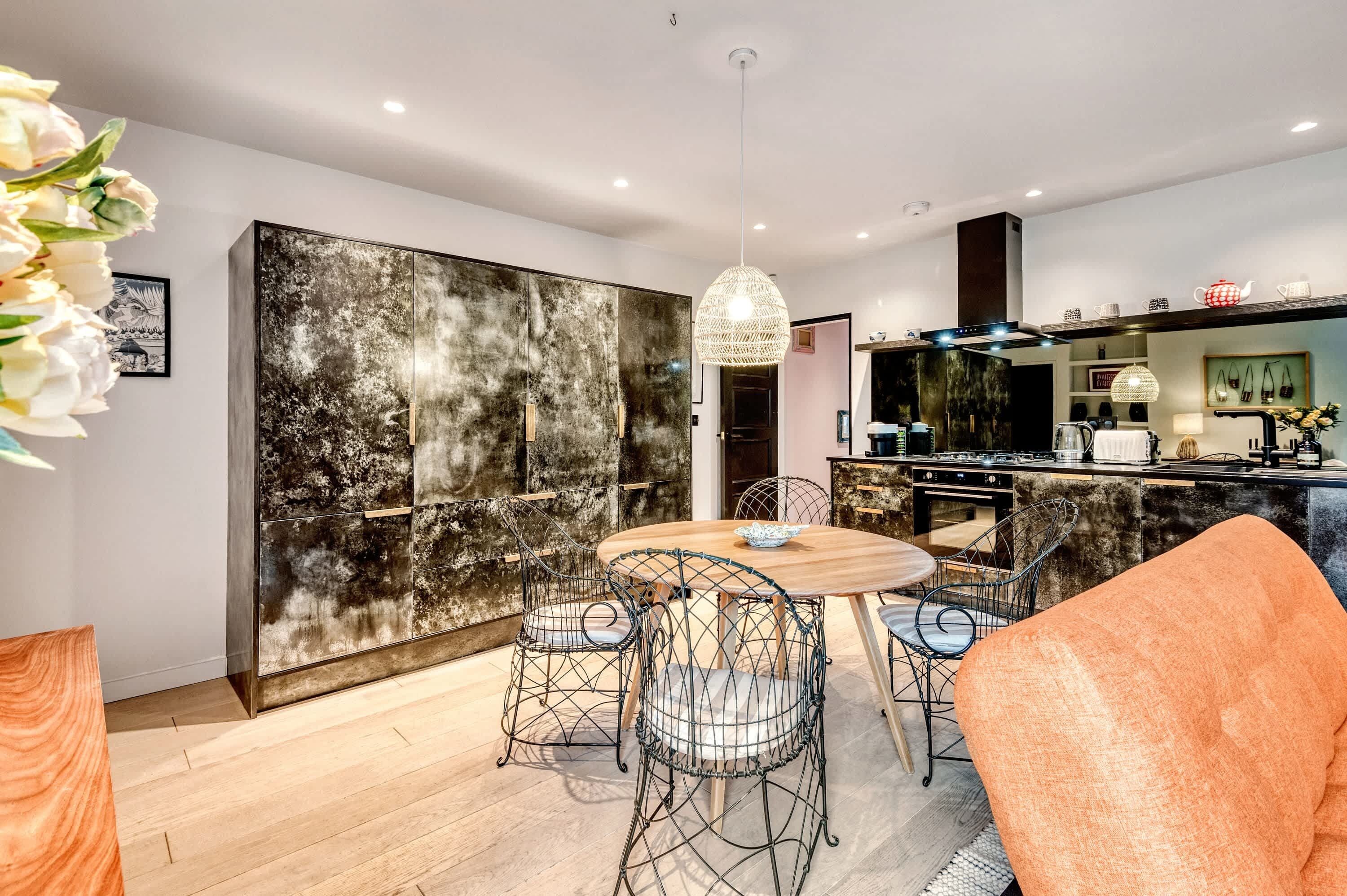 A modern kitchen with a dining area, featuring a round table with wire chairs, a hanging light fixture, and a large cabinet with a textured wall.