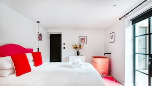 Un dormitorio moderno con una cama grande, un tocador blanco, un barril de madera y un ventanal amplio.