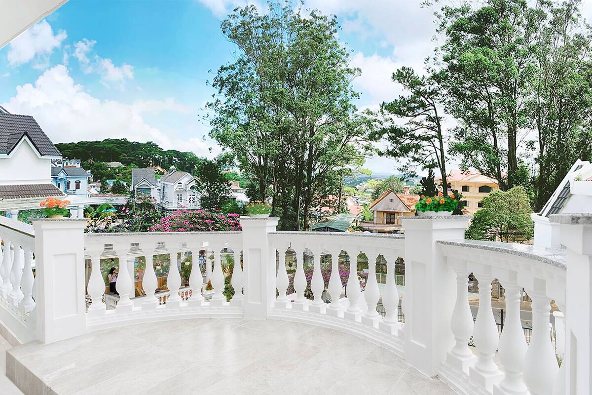 A balcony with a white balustrade, a tiled floor, and a view of surrounding buildings and greenery.