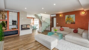 Un soggiorno moderno con divano, poggiapiedi e una scala in vetro che conduce a un balcone.