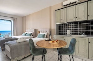 A hotel room with two beds, a dining table with chairs, a kitchen area, and a view of the sea.