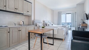 A modern kitchen with light wood cabinets, a dining table with a chair, a bed with white bedding, and a balcony with a view. - Studio Apartment 'Sunset Triple 1' with Sea View, Wi-Fi and Air Conditioning (LEFKADA)