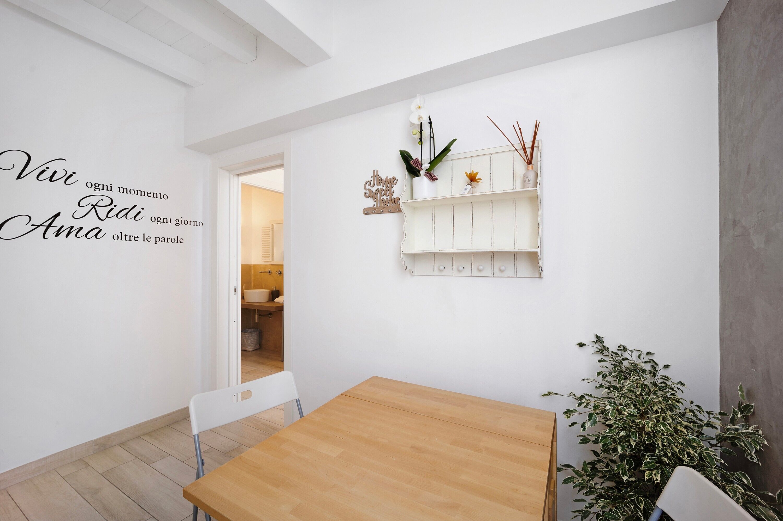 Un interno moderno con un tavolo in legno, sedie bianche, una parete bianca con testo motivazionale, uno scaffale bianco con oggetti decorativi e una pianta in vaso.
