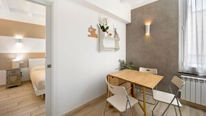 A room with a wooden dining table, white chairs, a white bed, a small white shelf with decorative items, and a wall-mounted light fixture. - 'Ama' Apartment with Balcony, Wi-Fi, and Air Conditioning (Albenga)