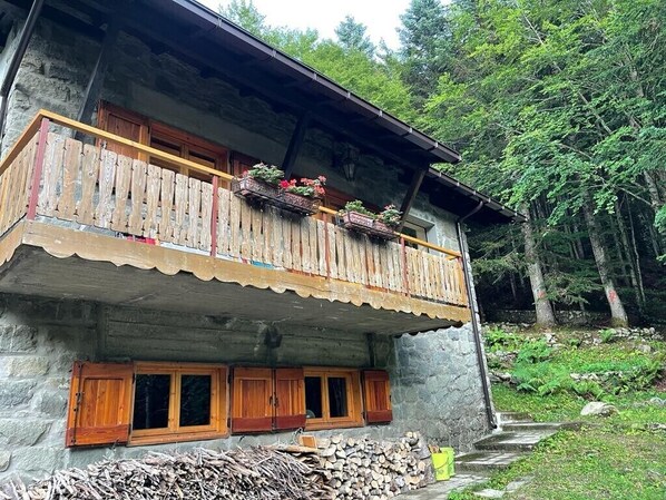 A stone building with a wooden balcony, windows with shutters, and a pile of firewood.