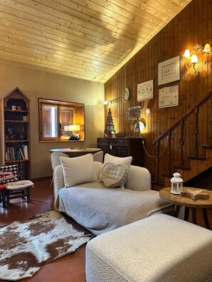 Een gezellige woonkamer met houten plafond, open haard, zithoek, boekenkast en een trap.