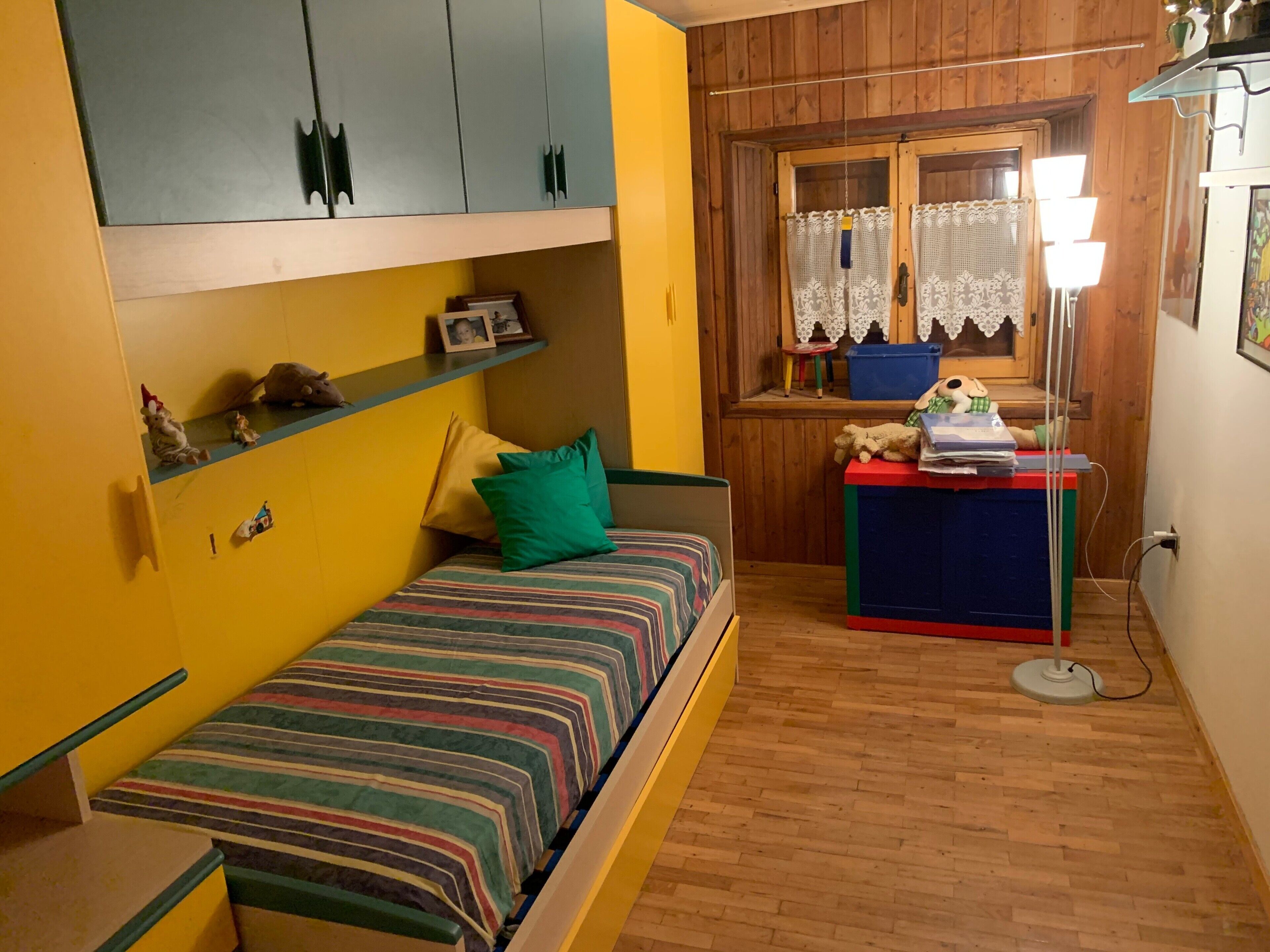 A small, cozy room with a single bed, a desk with a lamp, and a window with lace curtains.