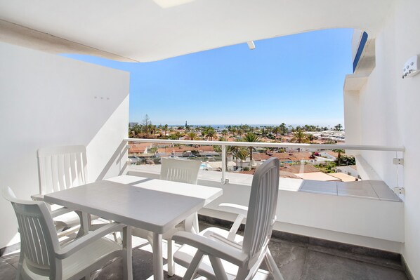 A balcony with a white table and chairs, a railing, and a view of a coastal town.