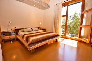 Une chambre avec un grand lit, une tête de lit en bois, des tables de chevet, une commode, une fenêtre avec des rideaux et une vue sur les arbres à l’extérieur.