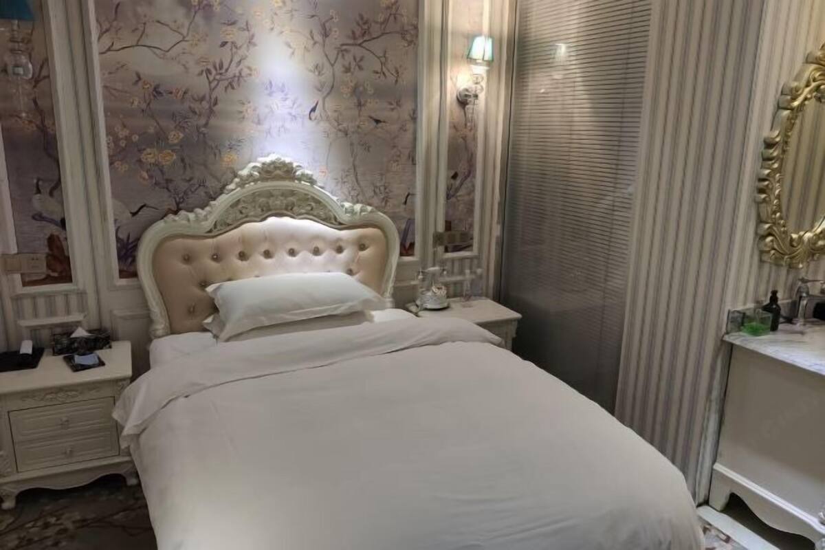 Ein Bett mit einer gepolsterten Kopflehne, ein Nachttisch mit einer Lampe und ein Spiegel an der Wand.
