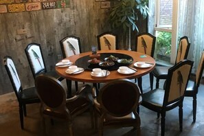 Une table de salle à manger ronde, entourée de chaises, avec des assiettes et un plat principal au centre, le tout placé devant un mur rustique agrémenté d’une fenêtre et d’une plante.