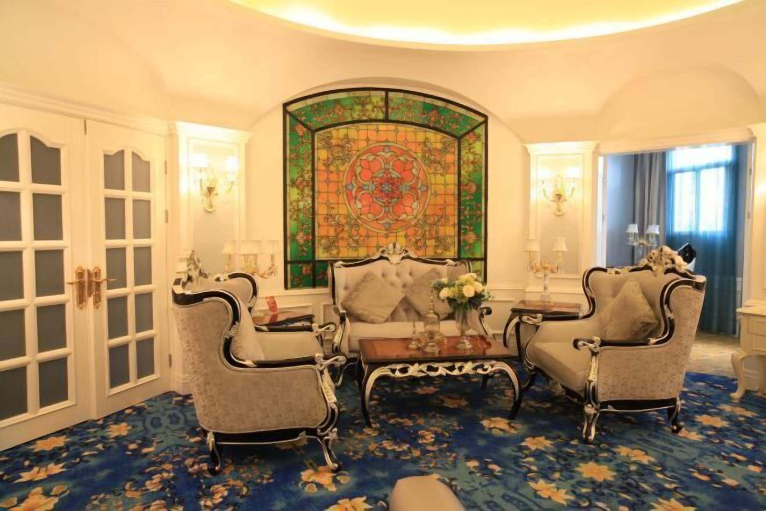 A room with a large stained glass window, two upholstered armchairs, a small table, and a patterned carpet.