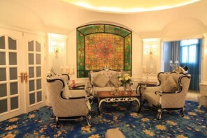 Um quarto com uma grande janela de vitrais, duas poltronas estofadas, uma mesinha e um tapete estampado.