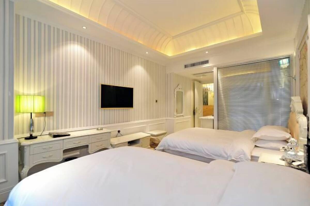 Ein Hotelzimmer mit zwei Betten, einem Fernseher, einem Schreibtisch mit Lampe und einem Fenster mit Jalousien.