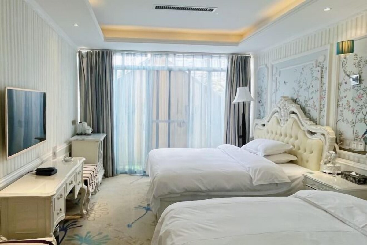 Une chambre d’hôtel avec deux lits, une grande fenêtre avec des rideaux, une télévision et un panneau décoratif au mur.