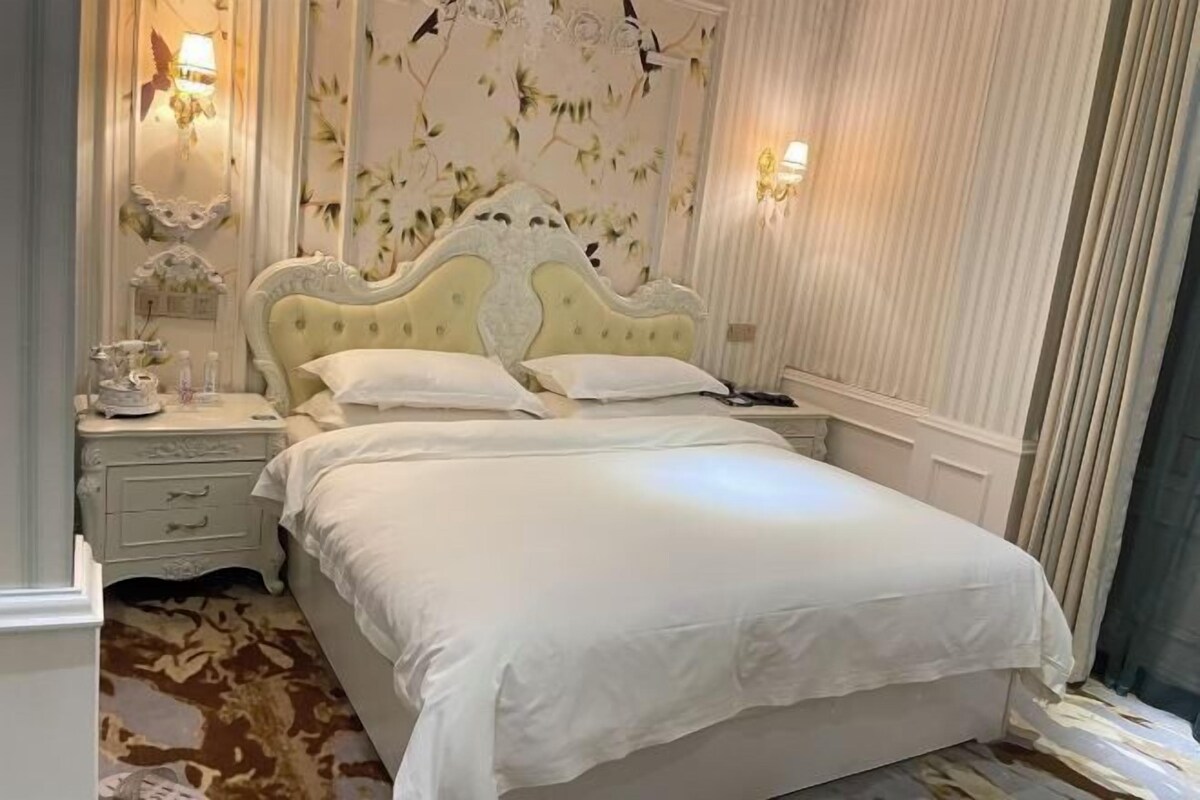 Un lit bien fait, recouvert d’une courtepointe blanche et agrémenté d’oreillers, une table de chevet avec une lampe, et un papier peint à motifs floraux sur le mur.