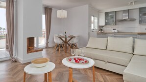 Un salon moderne comprenant un canapé, deux petites tables, un coin repas avec une table et des chaises, et une cuisine équipée de placards et d’un plan de travail.
