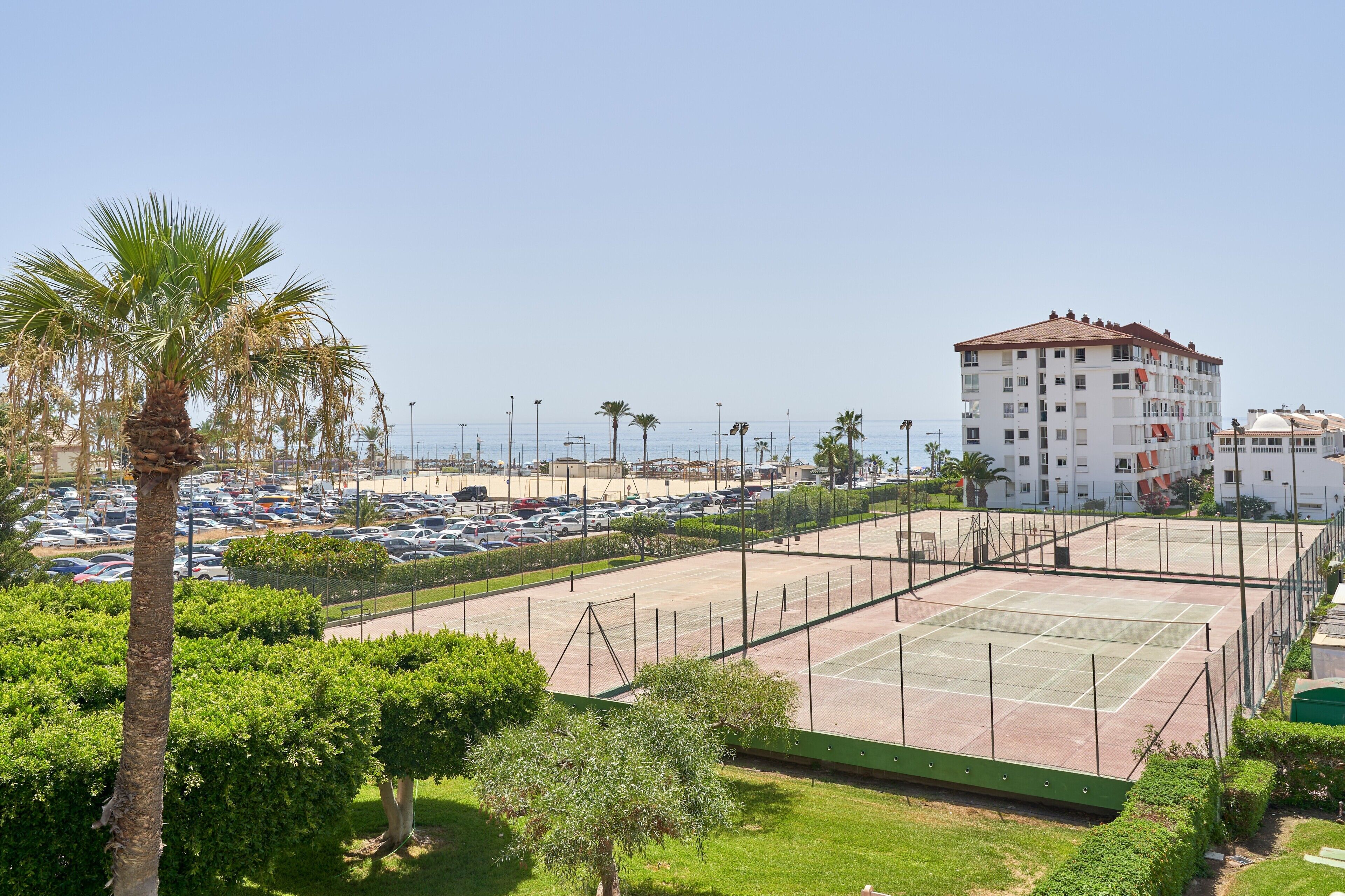 Una cancha de tenis rodeada de vegetación, un estacionamiento y un edificio de varios pisos con balcones.