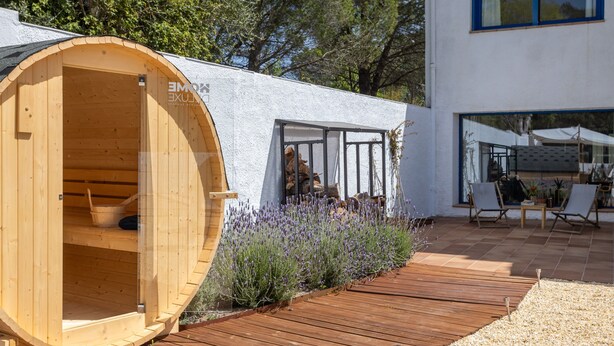 Eine holzgefässartige Konstruktion mit einer Tür, eine Terrassenfläche mit Holzboden und ein Gebäude mit großen Fenstern.