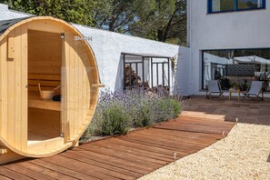 A wooden barrel-shaped structure with a door, a patio area with wooden decking, and a white building with large windows. - Holiday Home 'Mas Tinus - Lavanda' with Mountain View, Private Pool and Wi-Fi (Santa Cristina D'Aro)