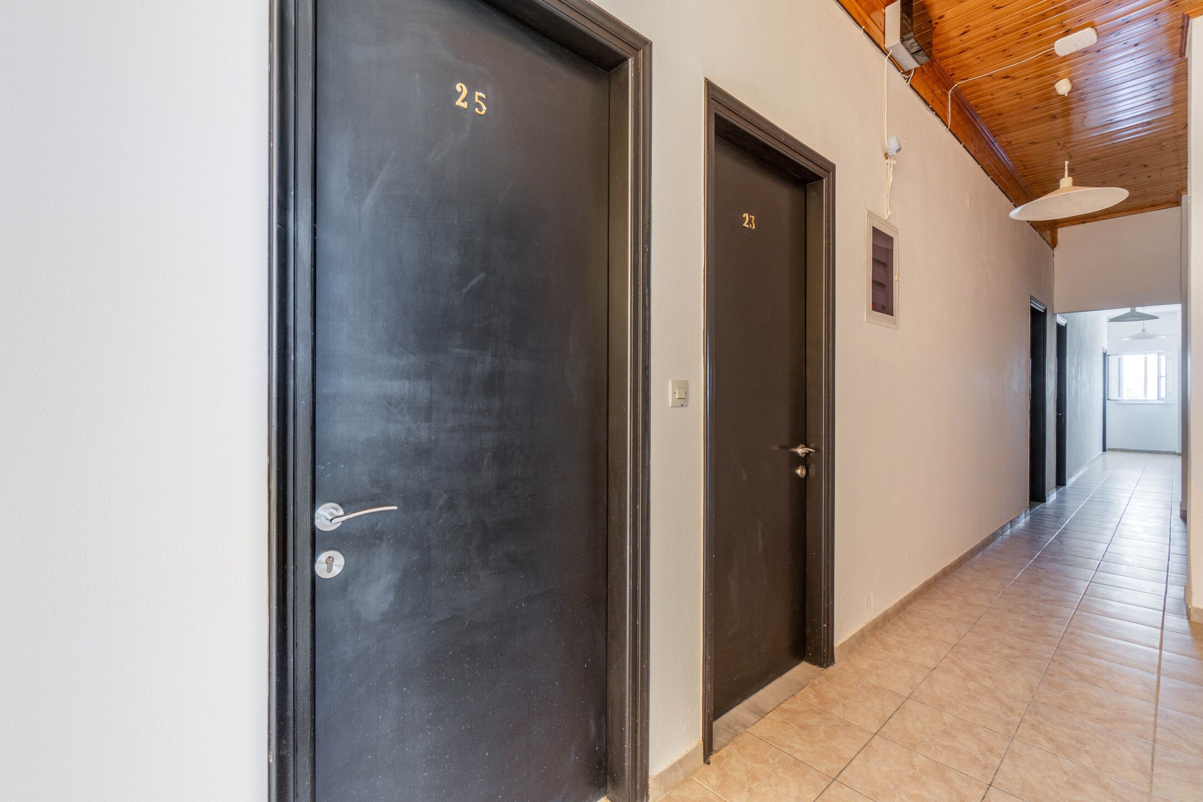 A hallway with two dark doors, numbered 25 and 23, leading to rooms. The hallway has a tiled floor and a wooden ceiling with exposed beams.