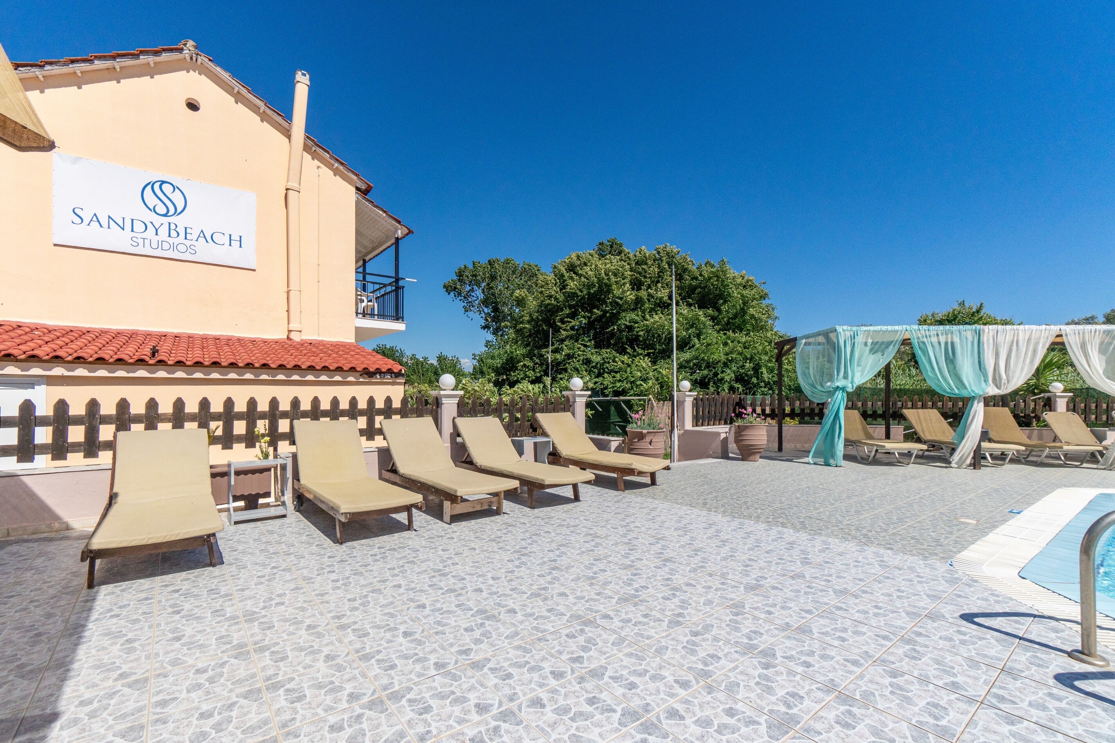 A pool area with lounge chairs, a building with a sign reading 'SANDYBEACH STUDIOS,' and a clear blue sky.