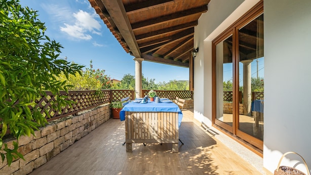 A covered patio with a wooden table and chairs, a stone wall, and a view of greenery.