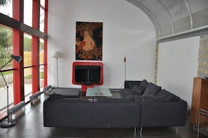 Un salon moderne avec un canapé d’angle, un téléviseur à écran plat et une grande fenêtre aux montants rouges.