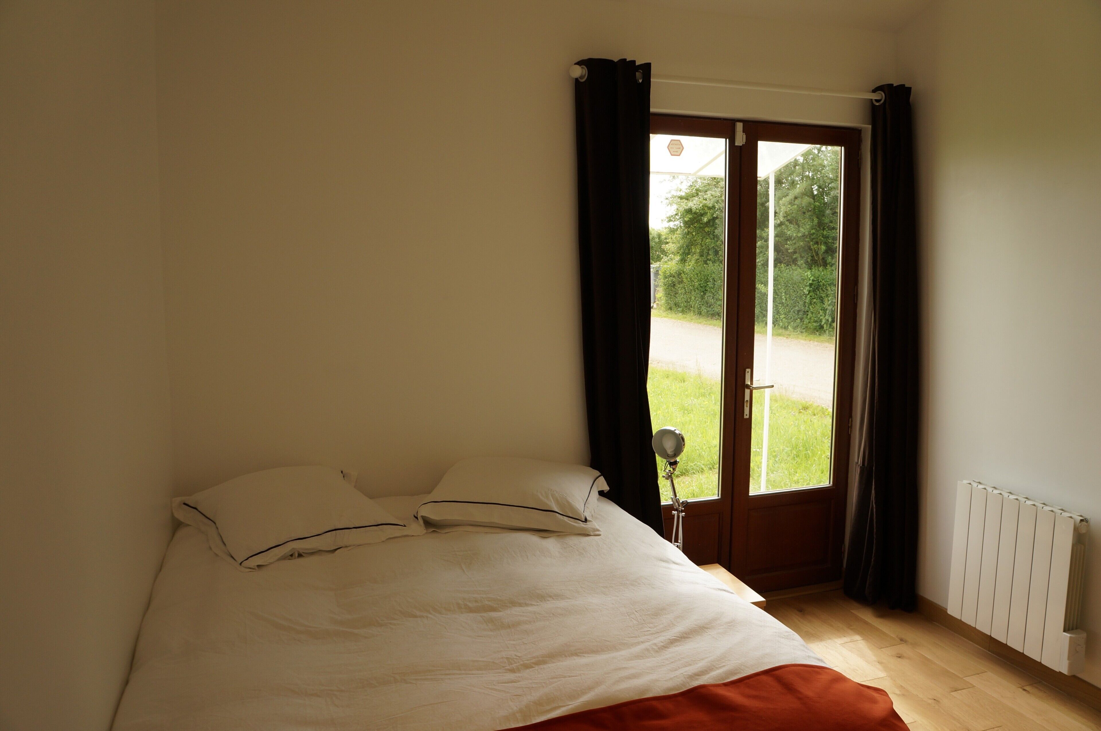 Une chambre à coucher avec un lit, une fenêtre avec des rideaux, un radiateur et une vue sur la verdure à l’extérieur.