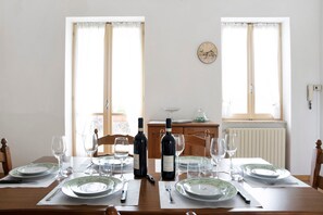 Mesa de jantar posta com pratos, talheres, copos de vinho e garrafas, com janelas e um relógio na parede.