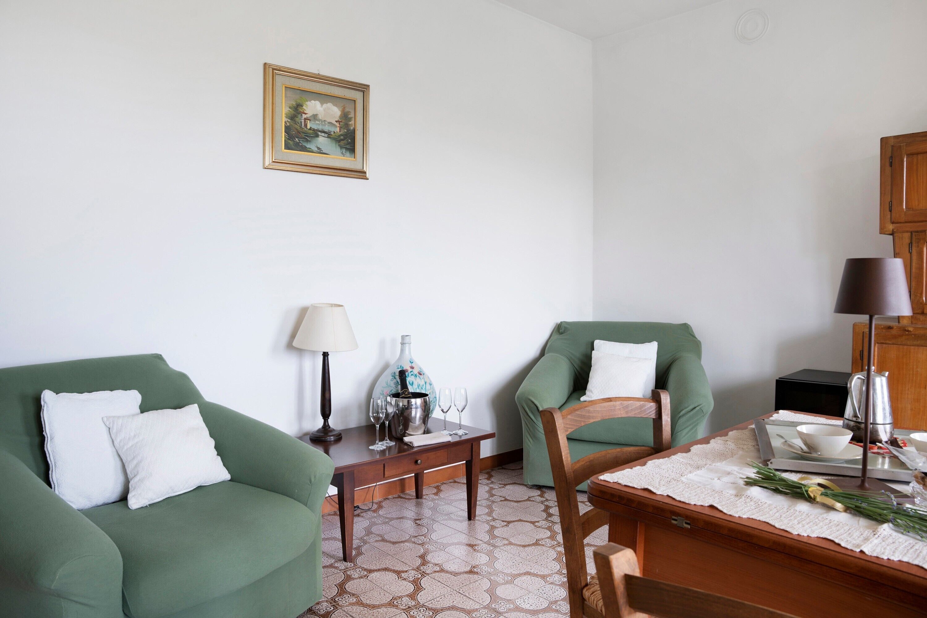 Un salon avec deux fauteuils verts, une table basse en bois, un vase blanc et un tableau encadré accroché au mur.