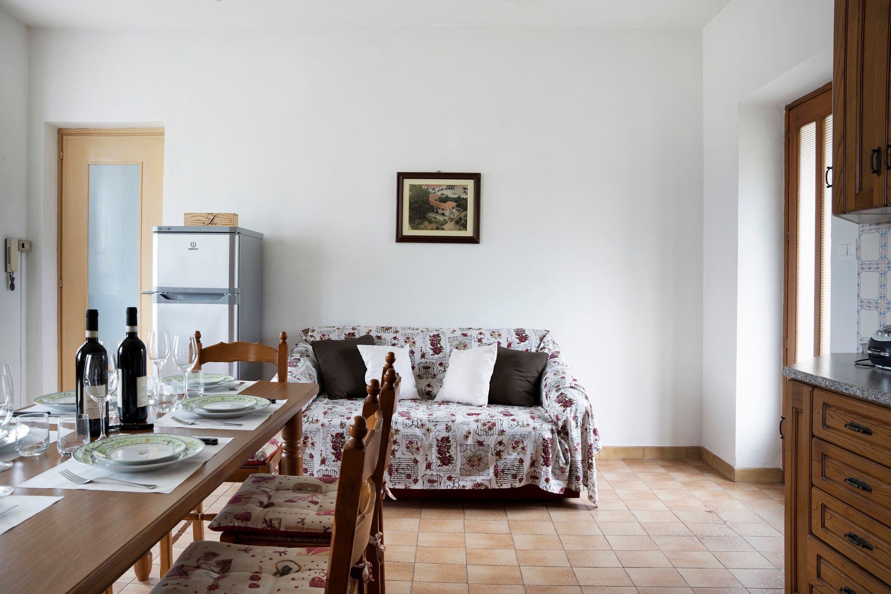 Un comedor con una mesa de madera dispuesta para cuatro, un sofá con tapicería floral, un cuadro en la pared y una zona de cocina con armarios de madera.