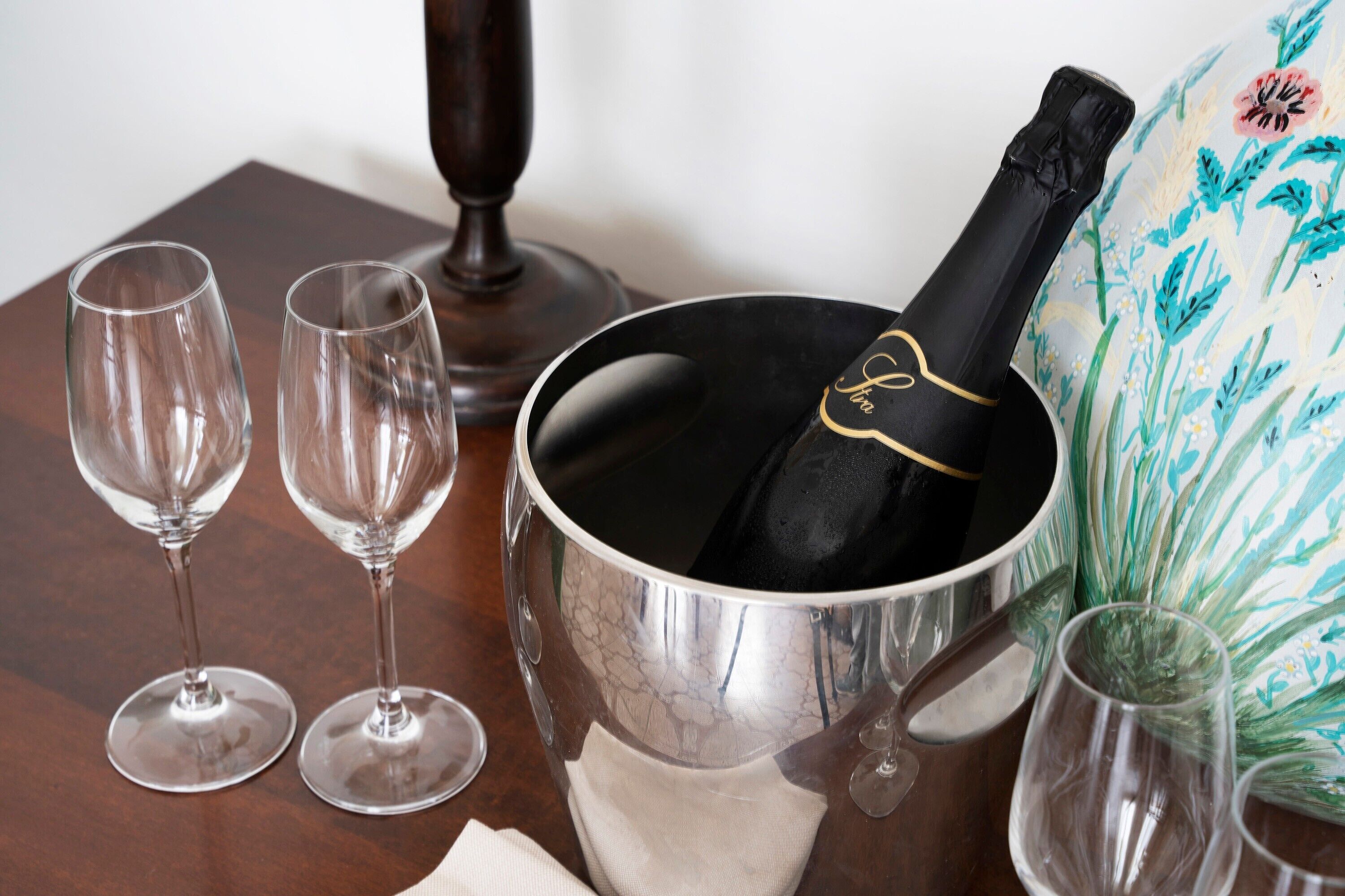 Ein Tisch mit einer Champagnerflasche in einem Metallkübel, zwei leere Weingläser und einem Dekorationsgegenstand mit floralem Muster.